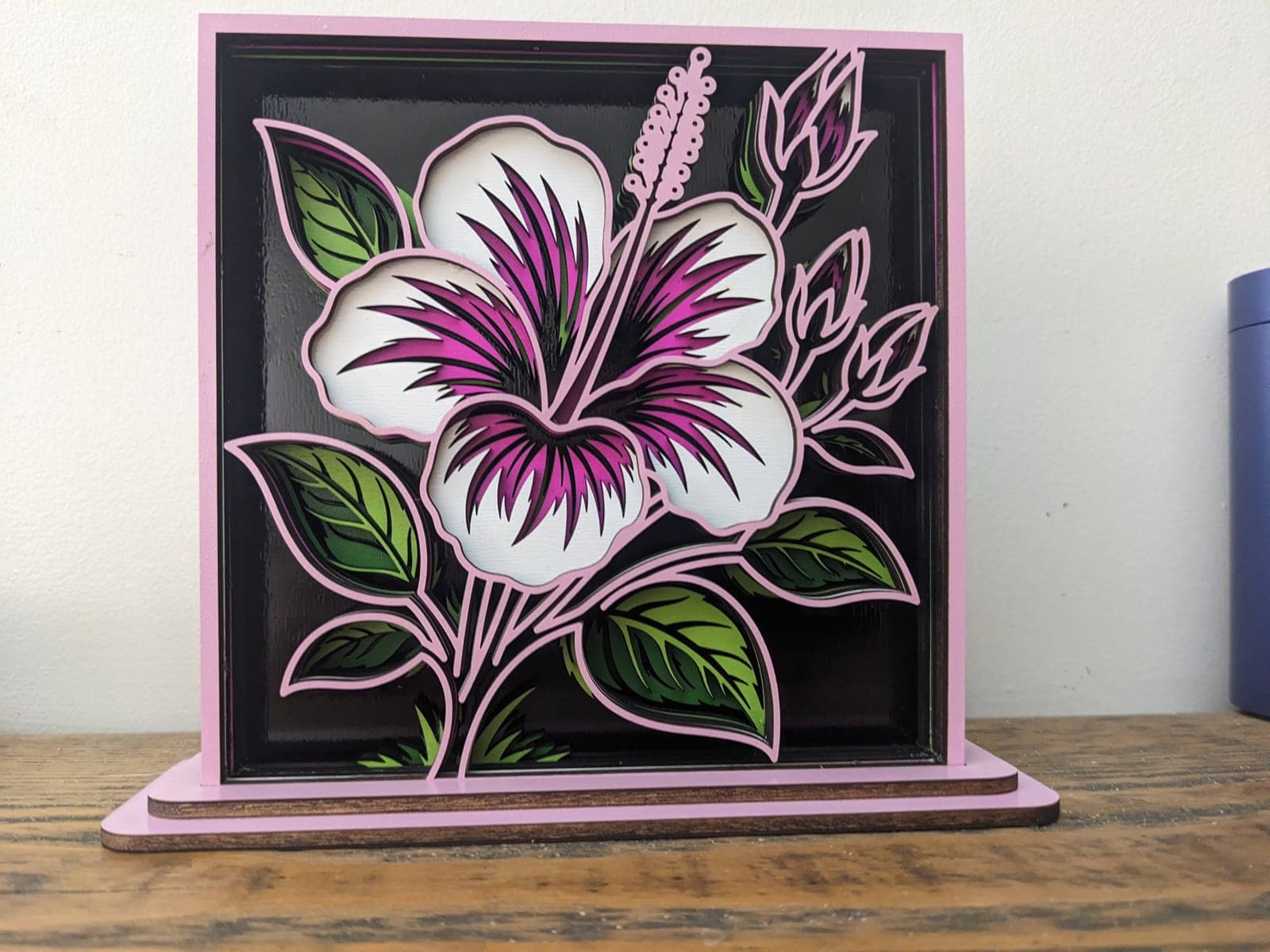 A layered wooden hibiscus flower in pink, white and green, in a pink stand